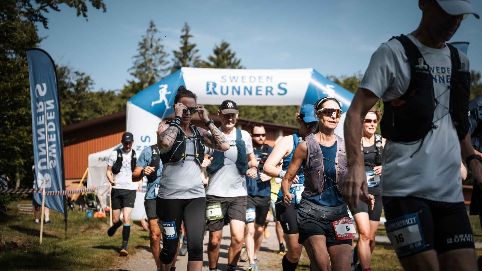 Grupp löpare från Sweden Runners startar ett lopp i Blekinge under en solig dag, iklädda löparvästar och sportutrustning, med en uppblåsbar målportal i bakgrunden och grönska runtomkring.