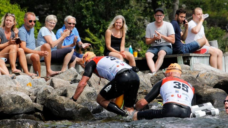 Två deltagare i swimrun klättrar upp på stenar från vattnet, iklädda nummerlappar och orangea kepsar, medan en engagerad publik sitter på en brygga och applåderar i Blekinges natursköna miljö.