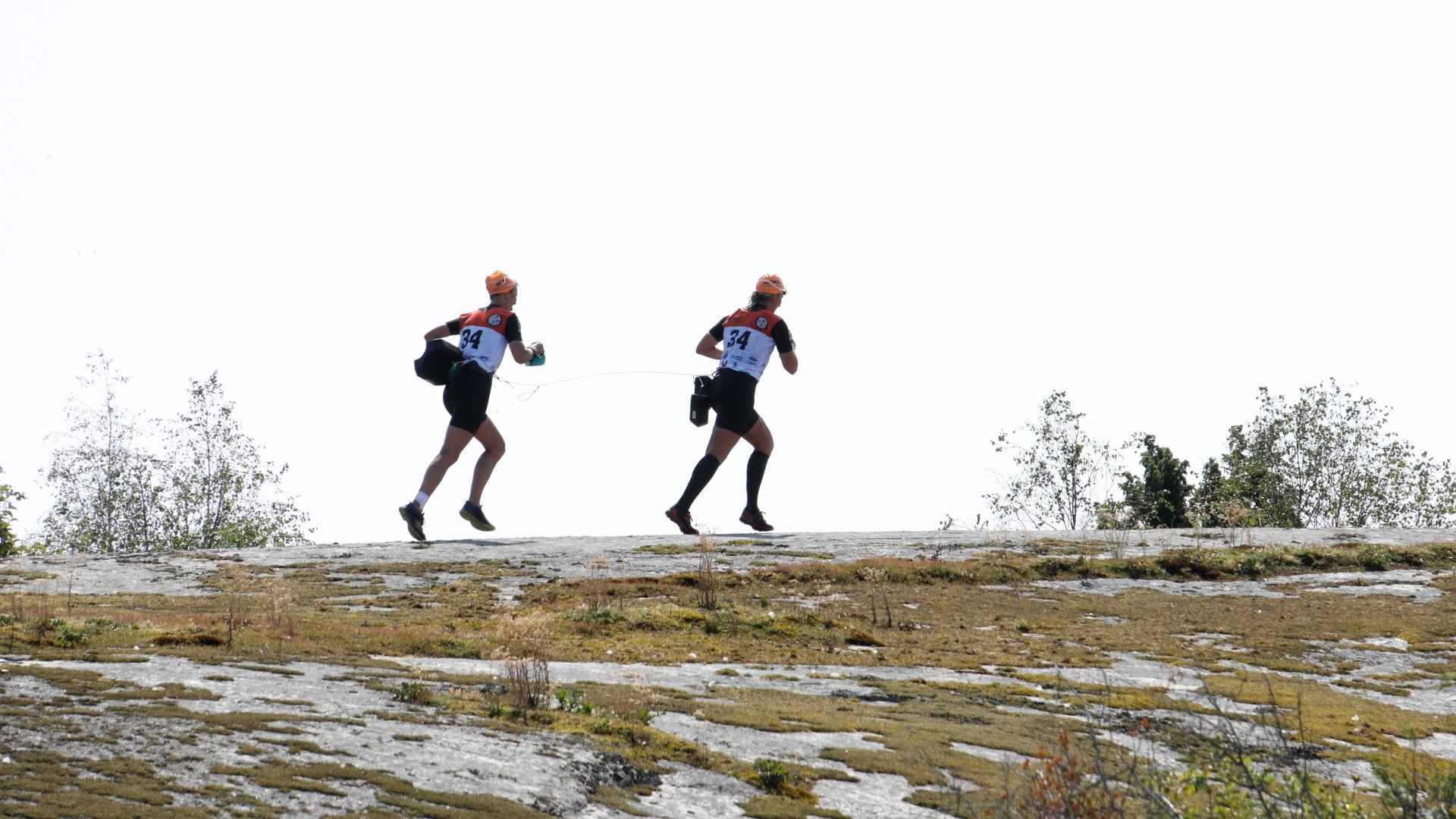 Två deltagare i swimrun, sammanlänkade med en lina, springer över en karg klippa med mossväxtlighet i Blekinge, iklädda nummerlappar och orangea kepsar.