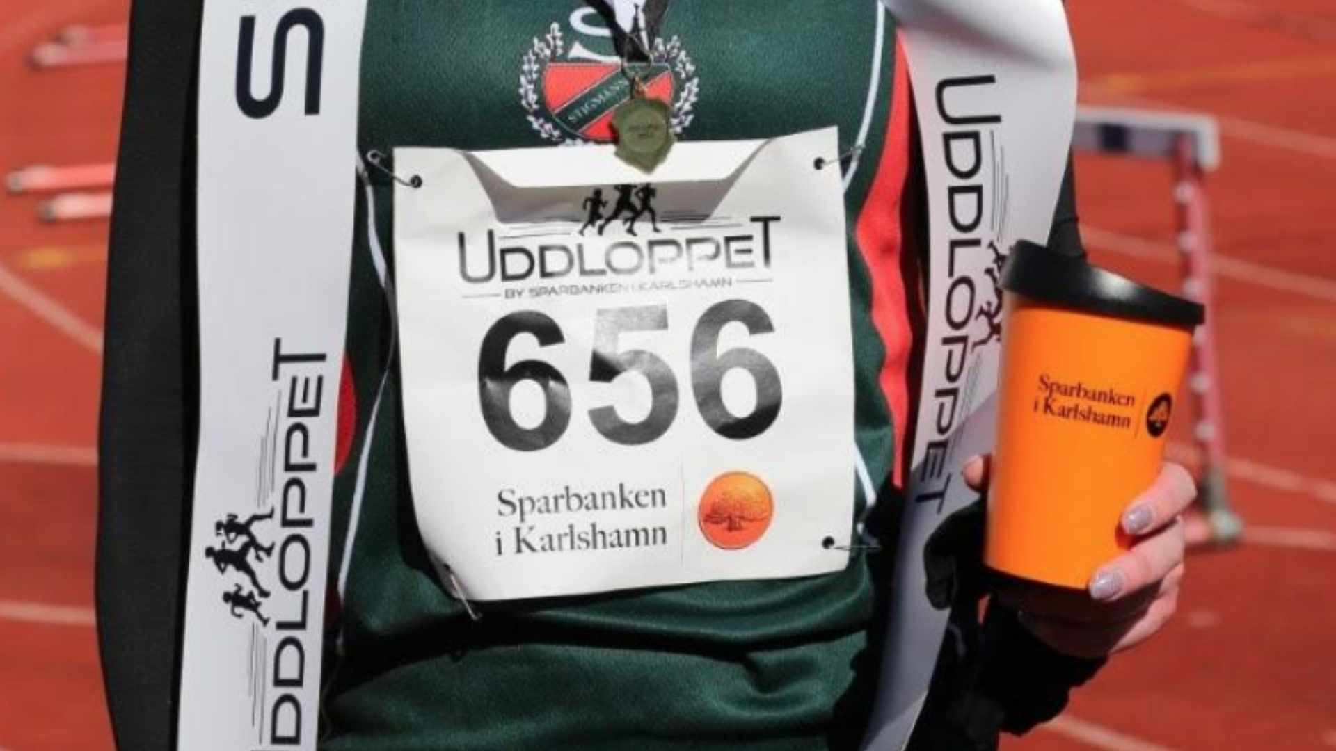 Närbild av en deltagare i Uddloppet med nummerlapp 656, iklädd grön tröja och medalj, hållandes en orange mugg från Sparbanken i Karlshamn på en löparbana i Blekinge.