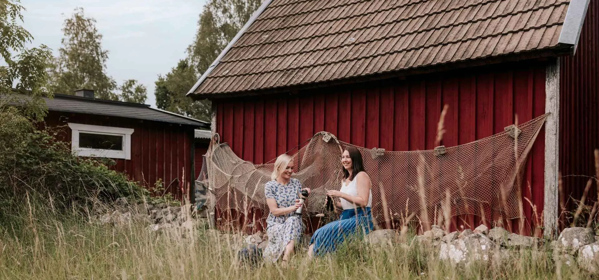 Två kvinnor njuter av våren framför en klassisk röd stuga i Blekinge, Sverige, omgivna av högt gräs och fisknät. Naturnära upplevelse i skärgårdsidyllen med svensk lantlig charm.