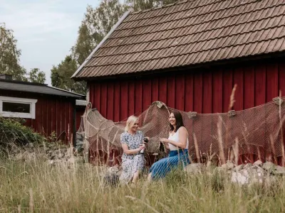 Två kvinnor njuter av våren framför en klassisk röd stuga i Blekinge, Sverige, omgivna av högt gräs och fisknät. Naturnära upplevelse i skärgårdsidyllen med svensk lantlig charm.